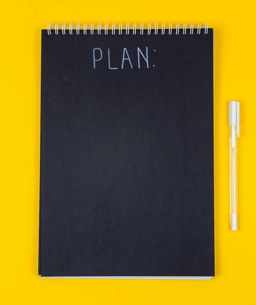 Schwarzer Ringblock und weißer Stift vor gelbem Hintergrund. Aufschrift „Plan“.
